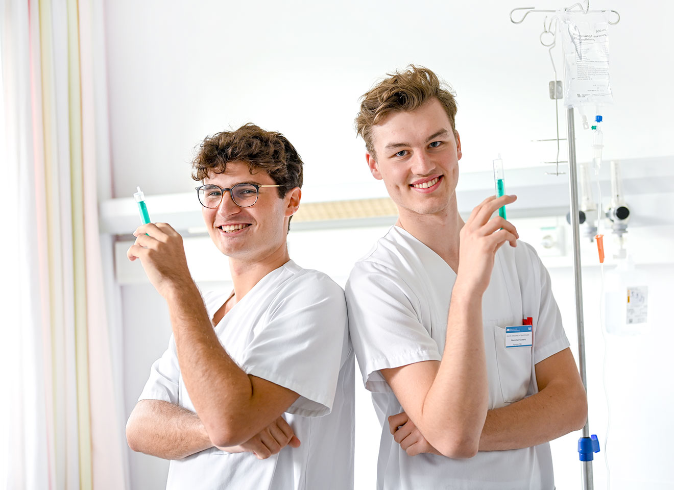 Zwei Auszubildende in der Pflege halten eine Spritze in der Hand und blicken in dir Kamera