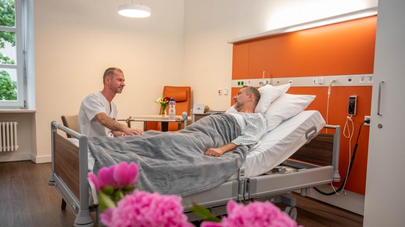 Patientenzimmer mit Patienten im Bett liegend und Pfleger am Bett sitzend Patientenzimmer mit Patienten im Bett liegend und Pfleger am Bett sitzend