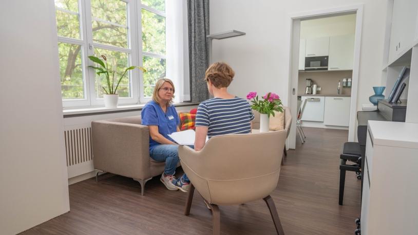 Blick in das Wohnzimmer mit Pflegerin auf Sofa und Patientin auf Stuhl sitzend 